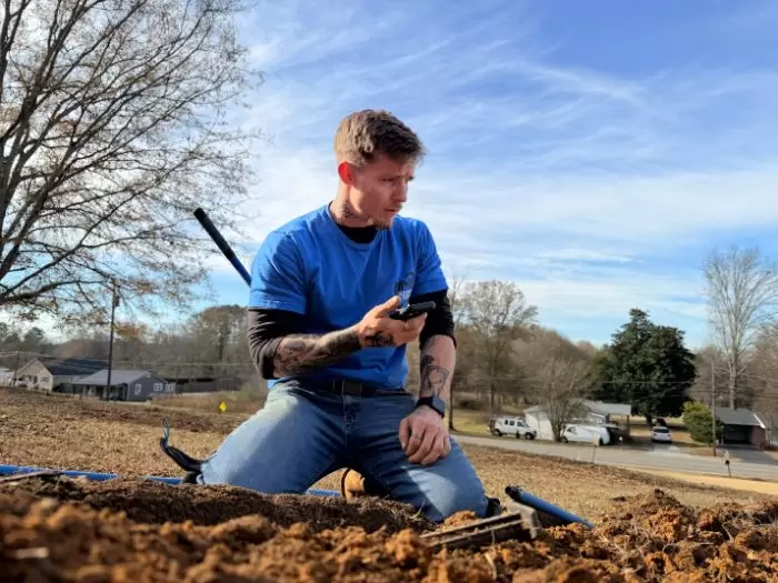 Plumber Checking Blue Water Line Trench With Phone Kneeling Yard